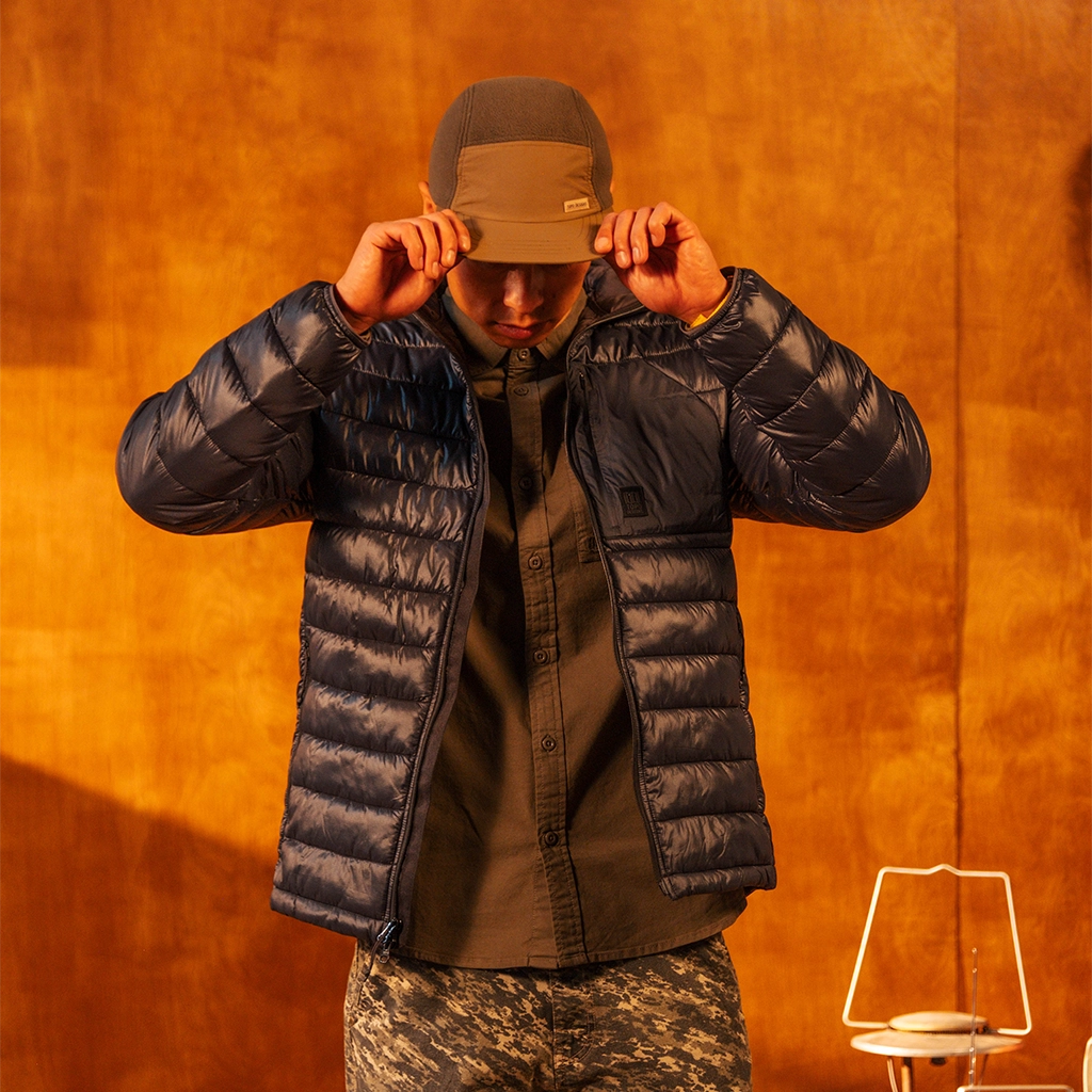 Close up of a person wearing a black puffer jacket and brown hat against an orange wall