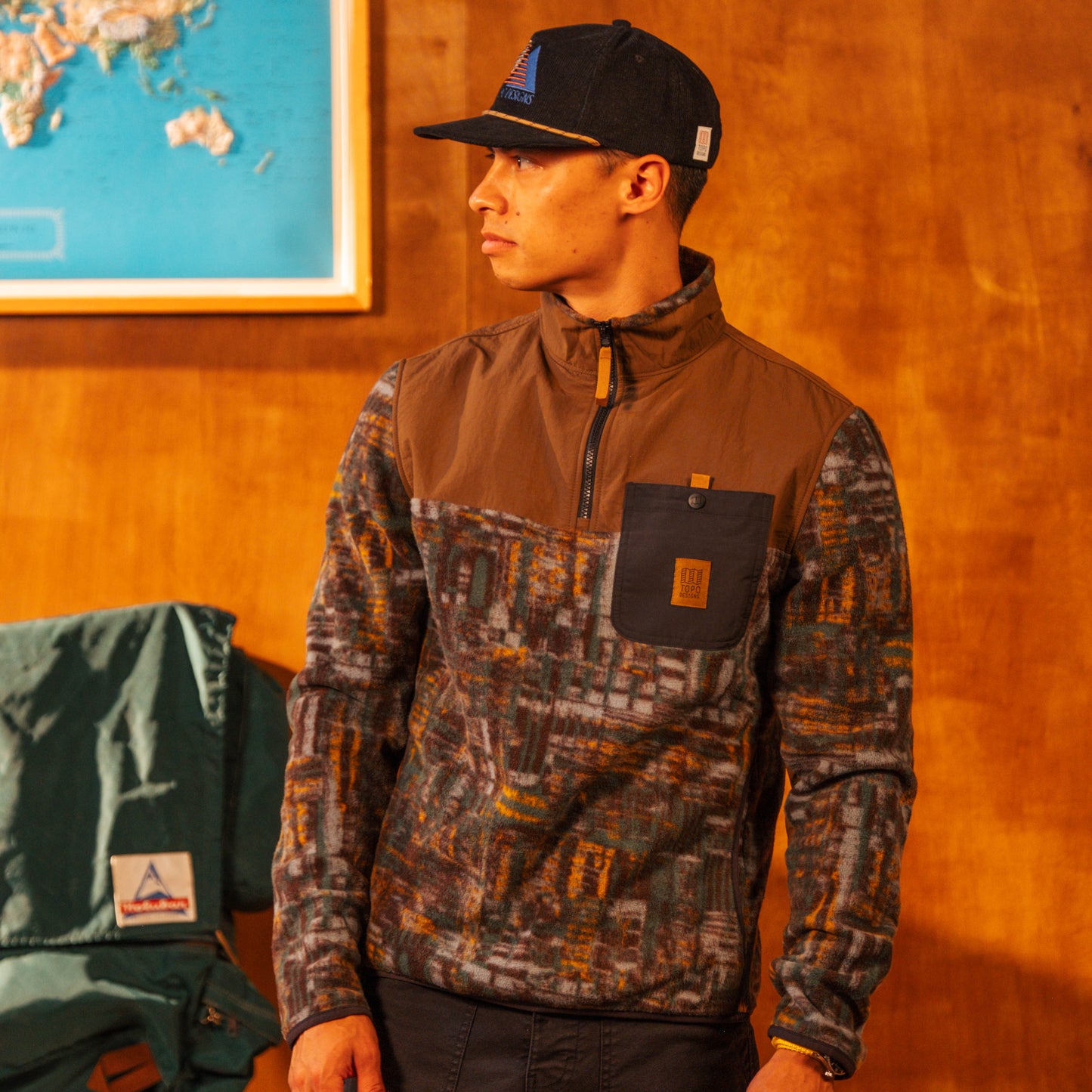 Man wearing a camouflage jacket and cap indoors with a map on the wall.