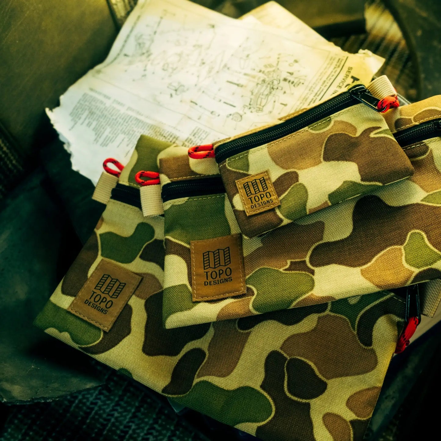 Three duck camouflage canvas pouches with black zippers and red pull tabs, each labeled "TOPO DESIGNS".