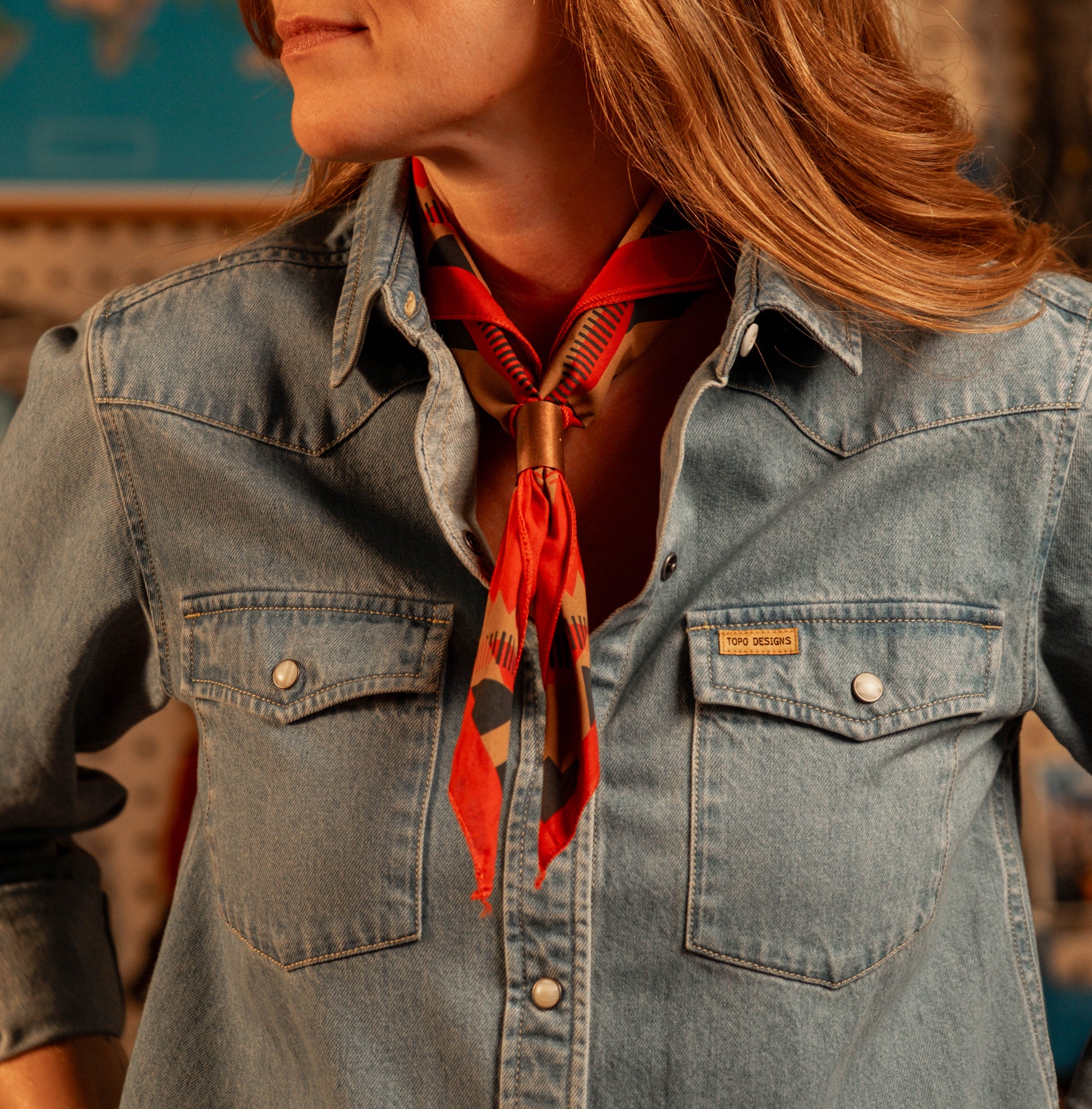 Person wearing a "light denim" shirt with a red patterned scarf around their neck, blurred background