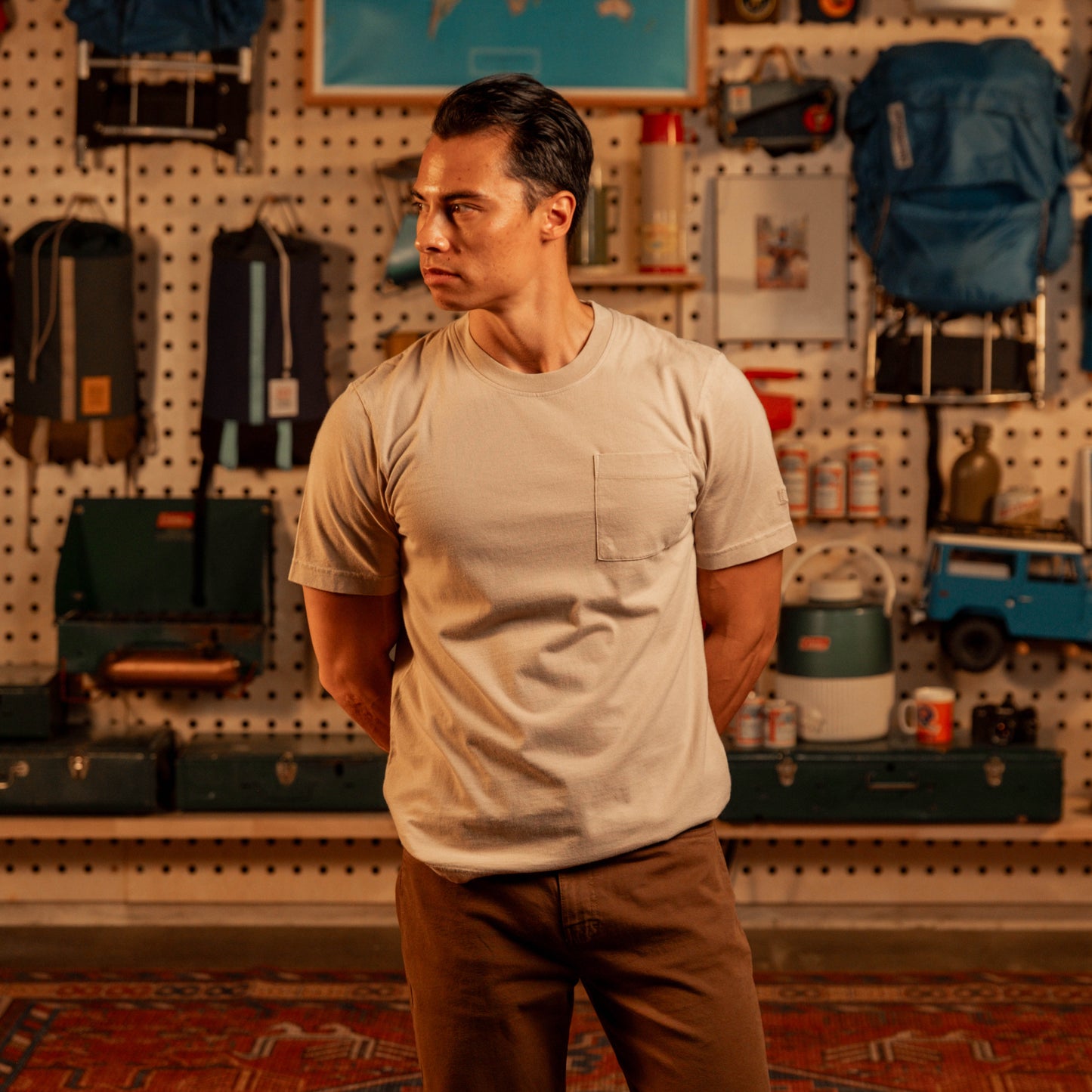 Man wearing off white t-shirt standing in front of wall with backpacks and camping gear.