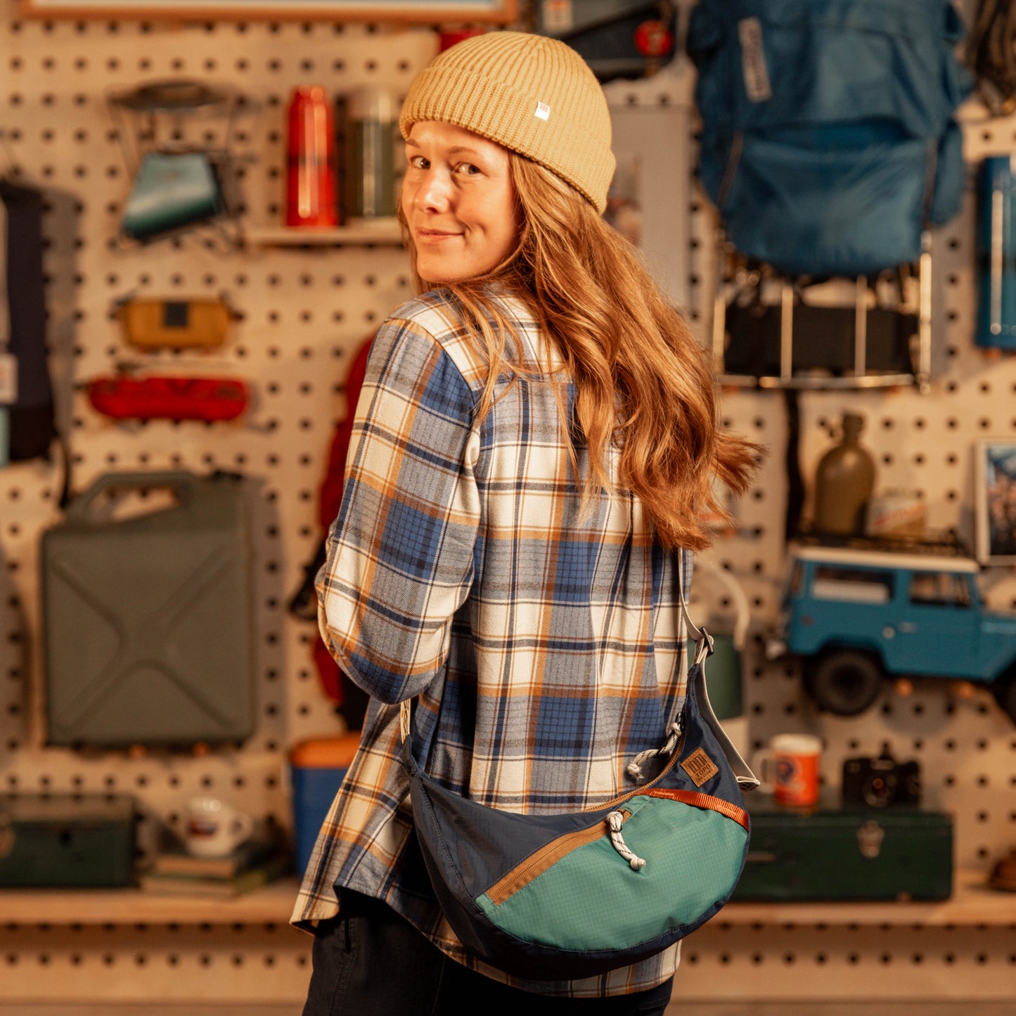 Person wearing a plaid shirt and beanie with a green bag, standing in a store with shelves and products.