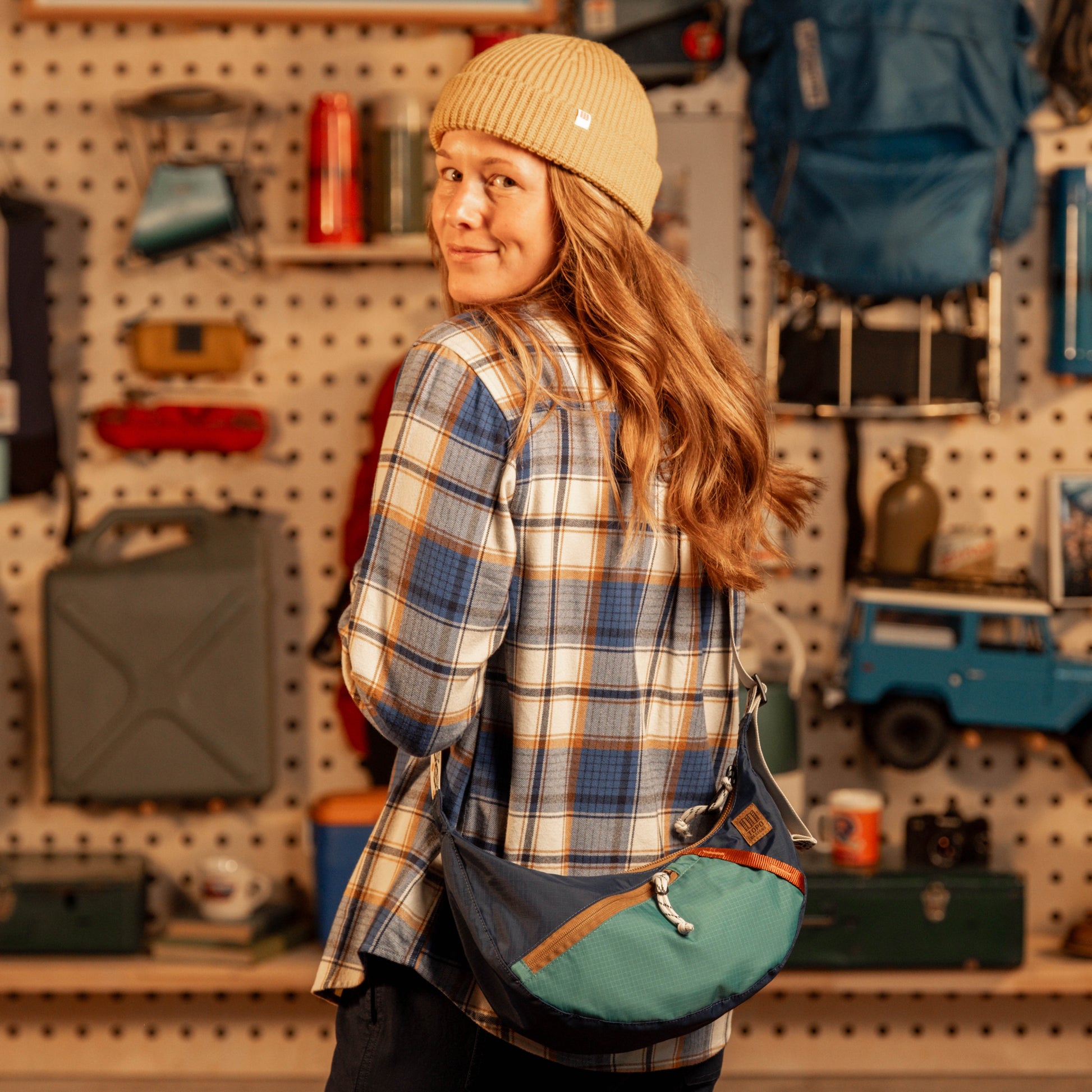 Person wearing a plaid shirt and beanie with a green bag, standing in a store with shelves and products.