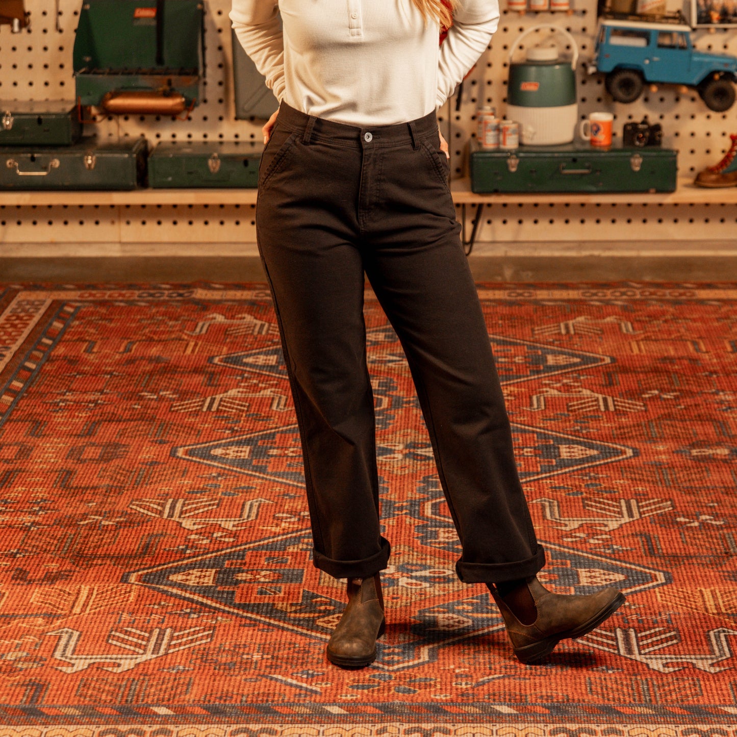 Person wearing dark pants and a white shirt standing on a patterned rug with a pegboard wall in the background.