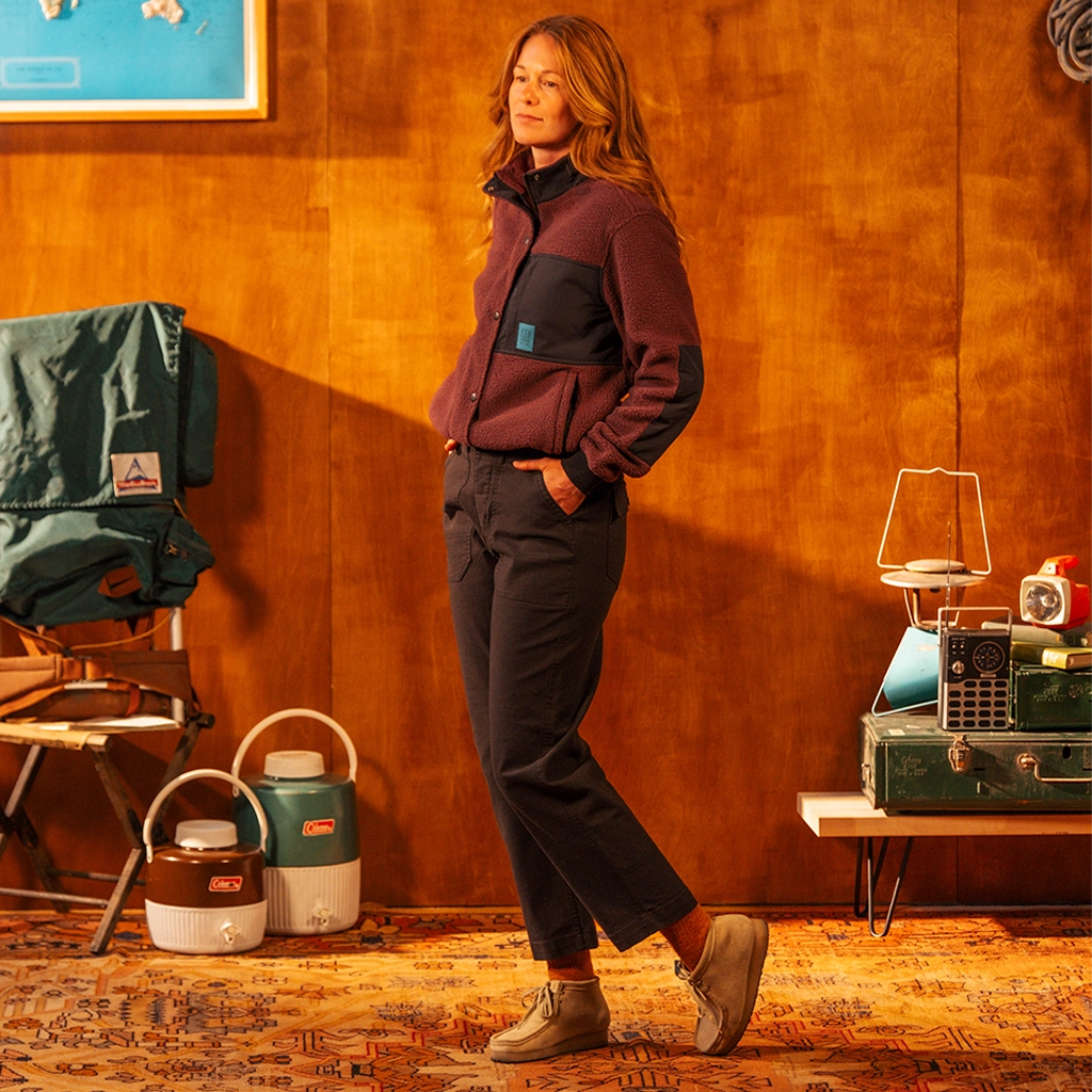 Person wearing a maroon jacket and dark pants standing in a room with camping gear