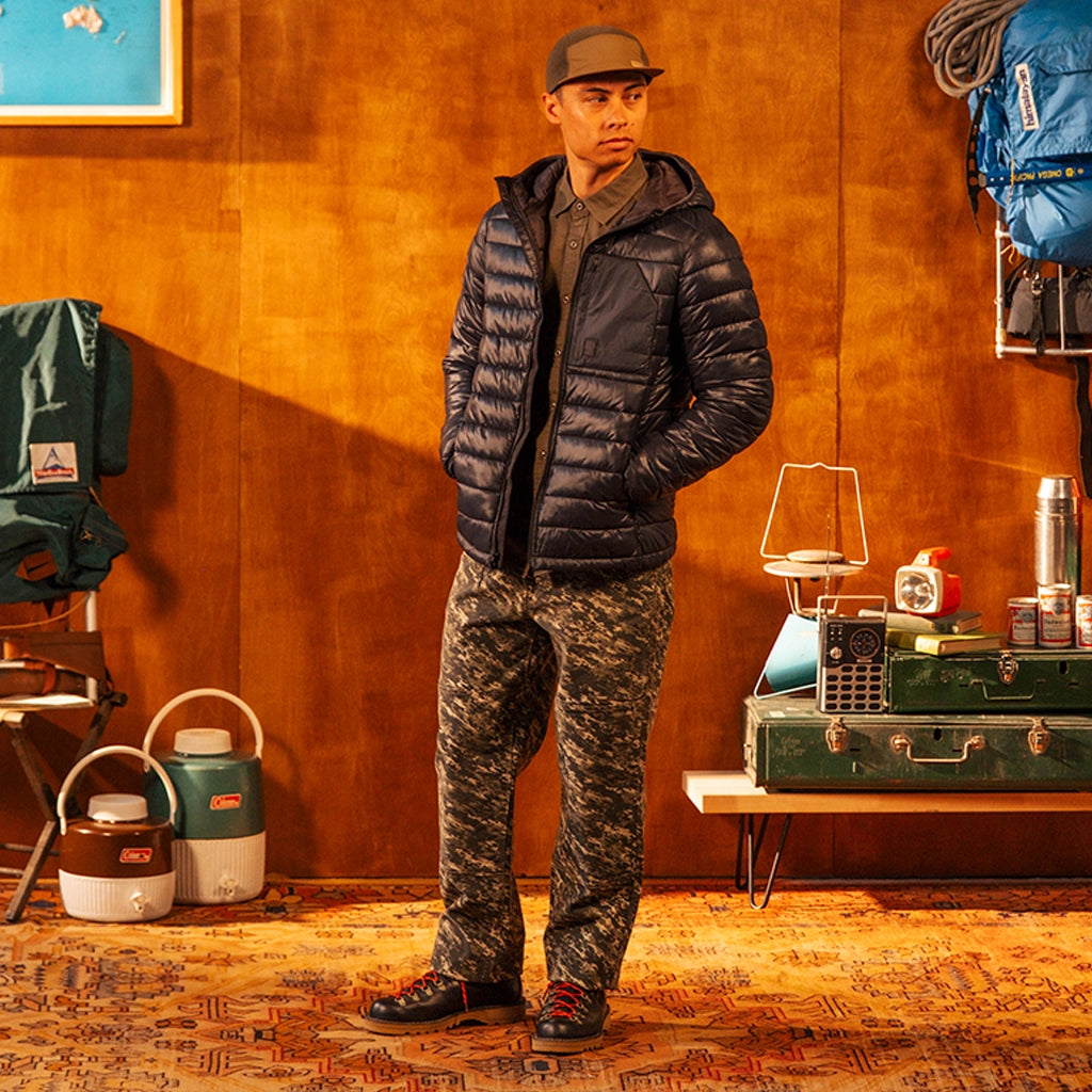 Person wearing a  black puffer jacket and brown hat against an orange wall 