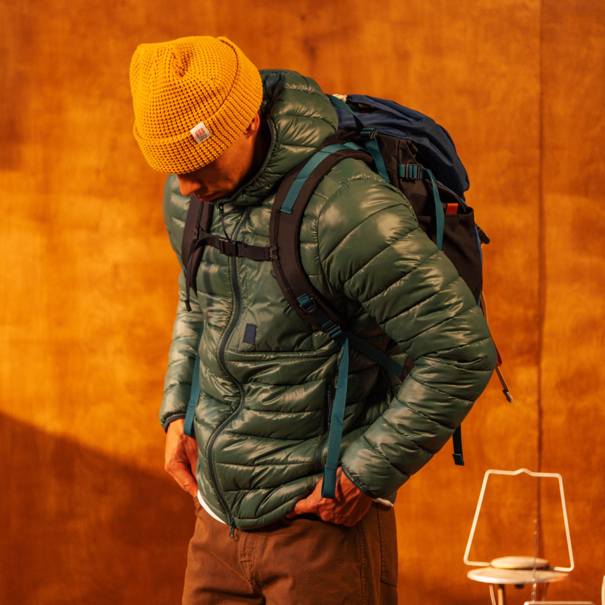 Person wearing a green puffer jacket and yellow beanie with a backpack against an orange wall