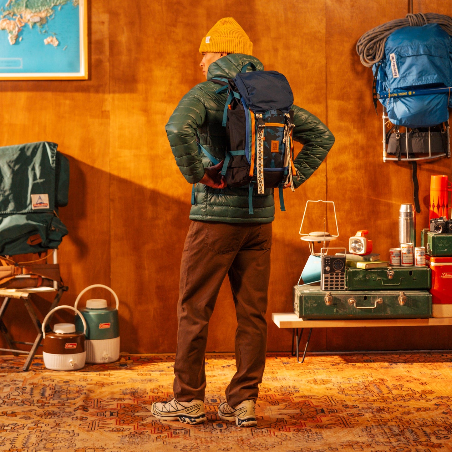 Person wearing a green jacket and backpack in a room with camping gear