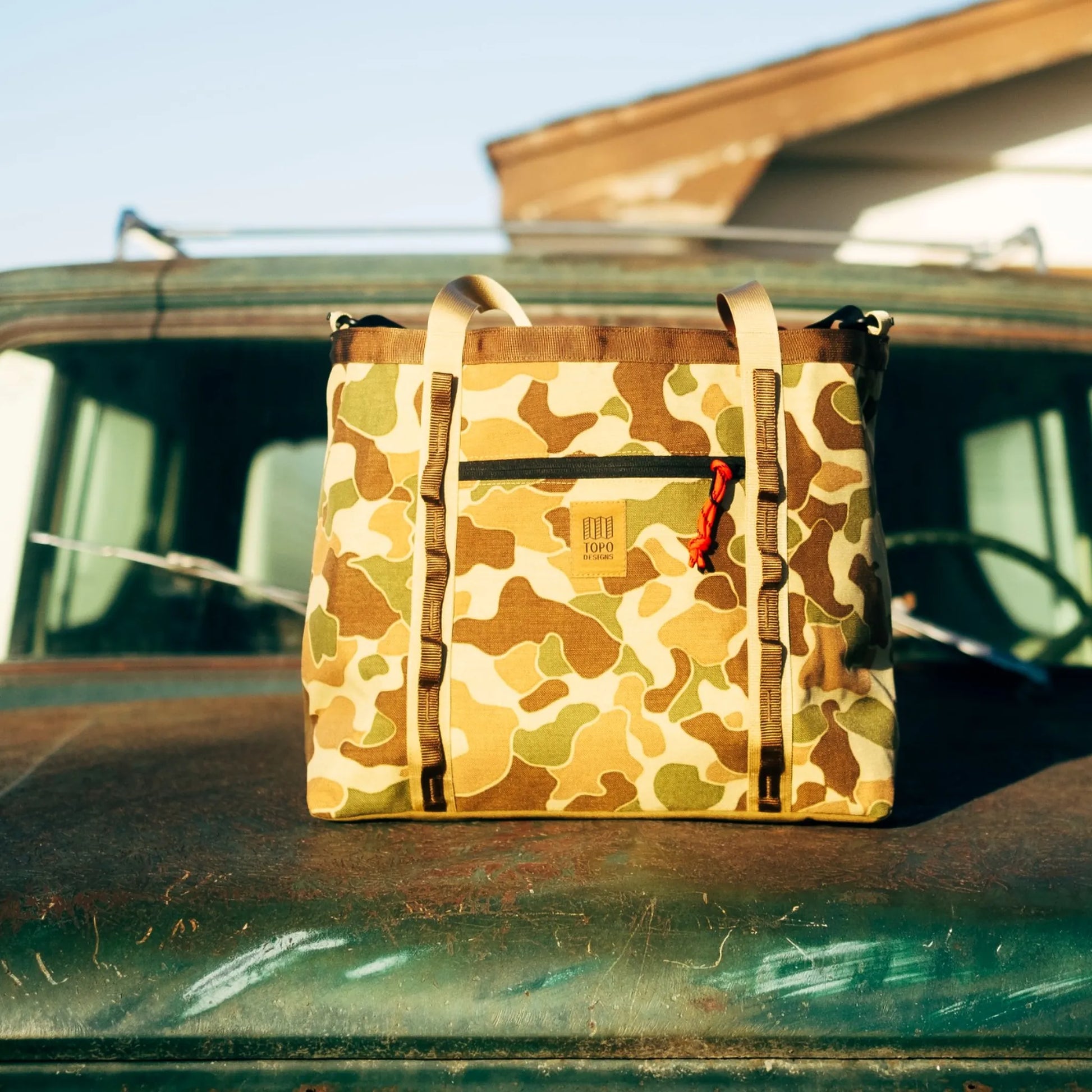 Duck camouflage tote bag with tan canvas handles and black zipper pocket, displayed on weathered green wood.