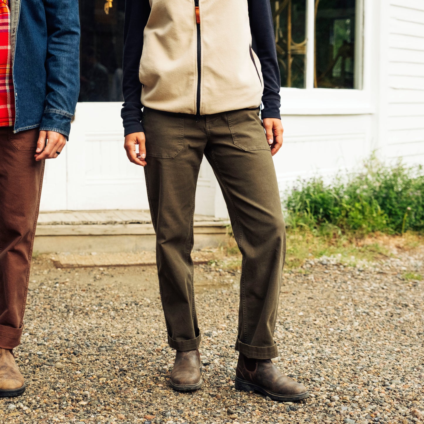 Two people wearing outdoor clothing standing on a gravel surface with a white building in the background.
