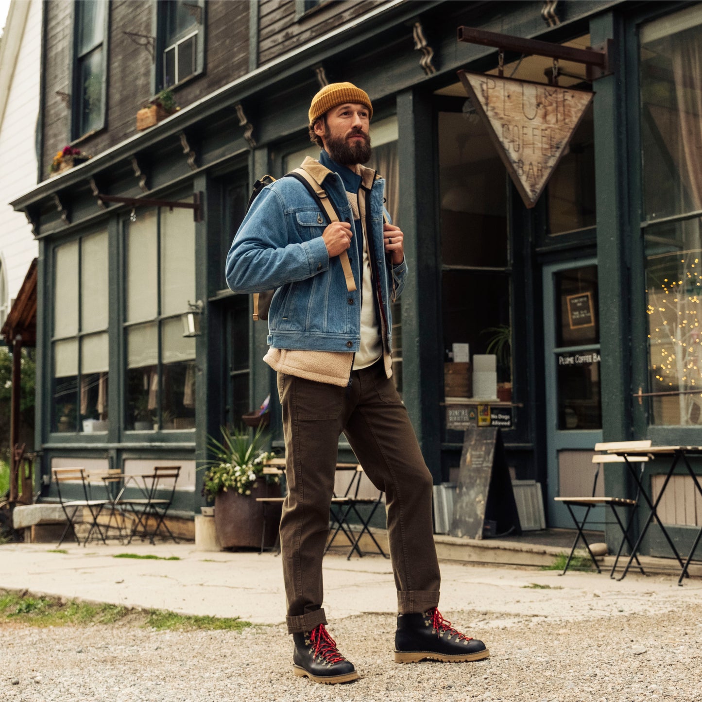 Man in a denim jacket and brown pants standing in front of a building with outdoor seating.