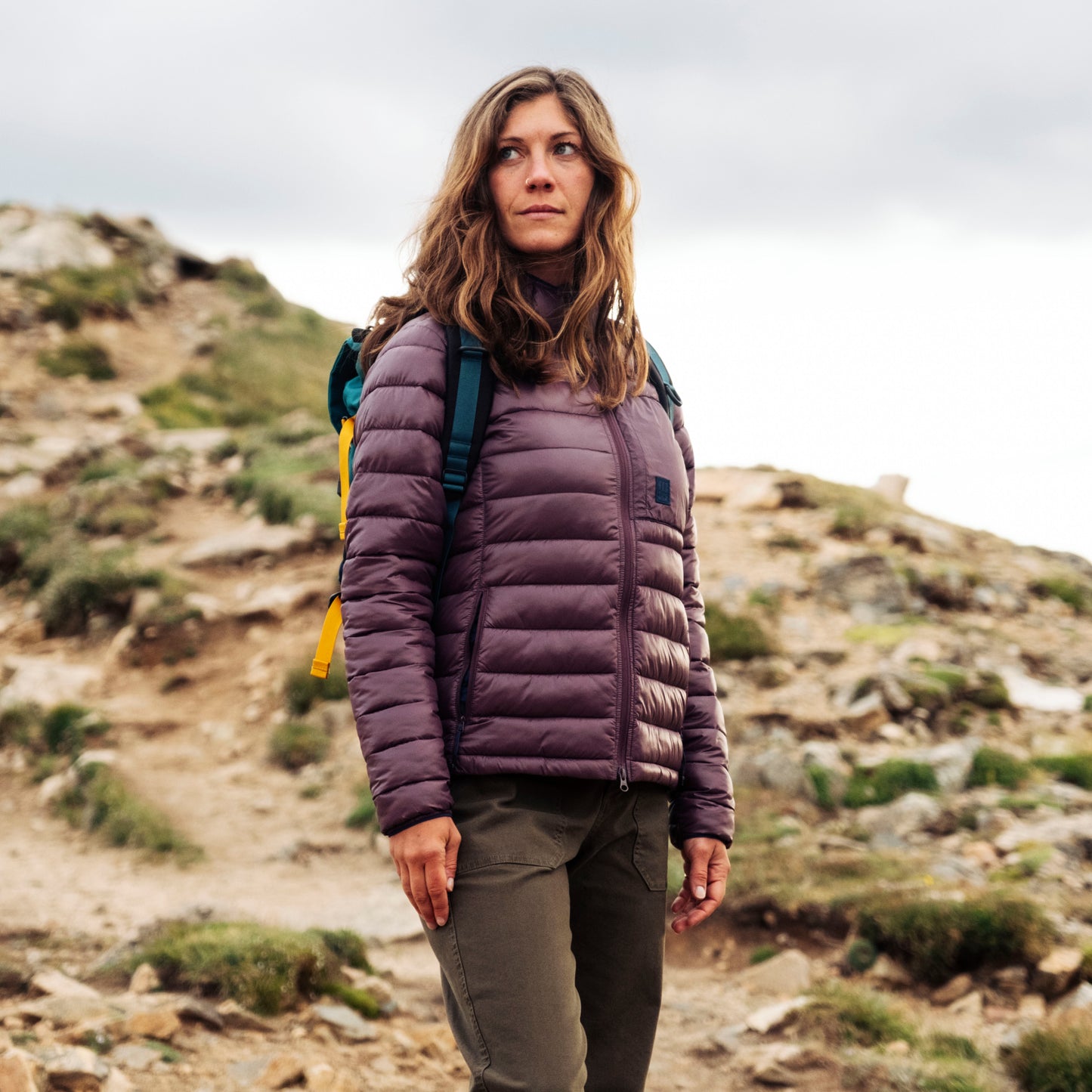 Woman in a purple jacket and green backpack hiking on a rocky mountain trail.