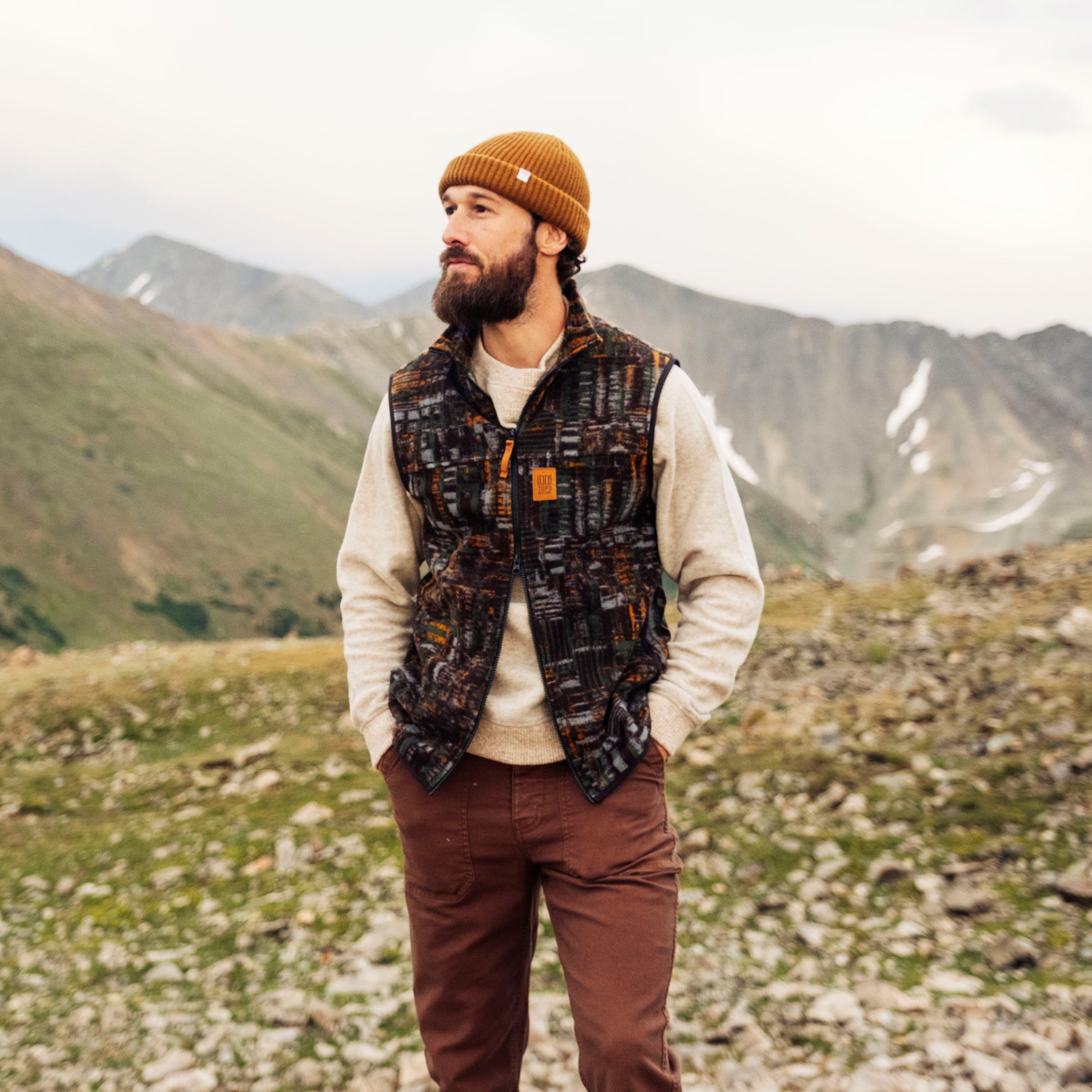 Man wearing a patterned vest and brown pants standing in a mountainous landscape.