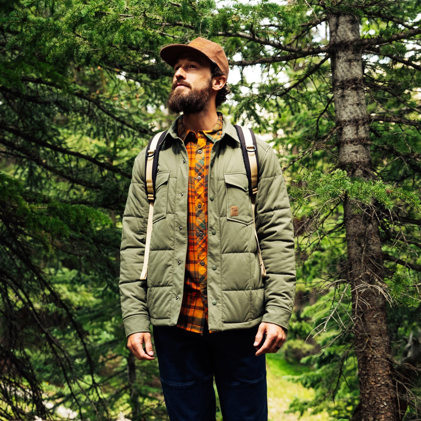Man in a forest wearing a green jacket and brown cap with a backpack.