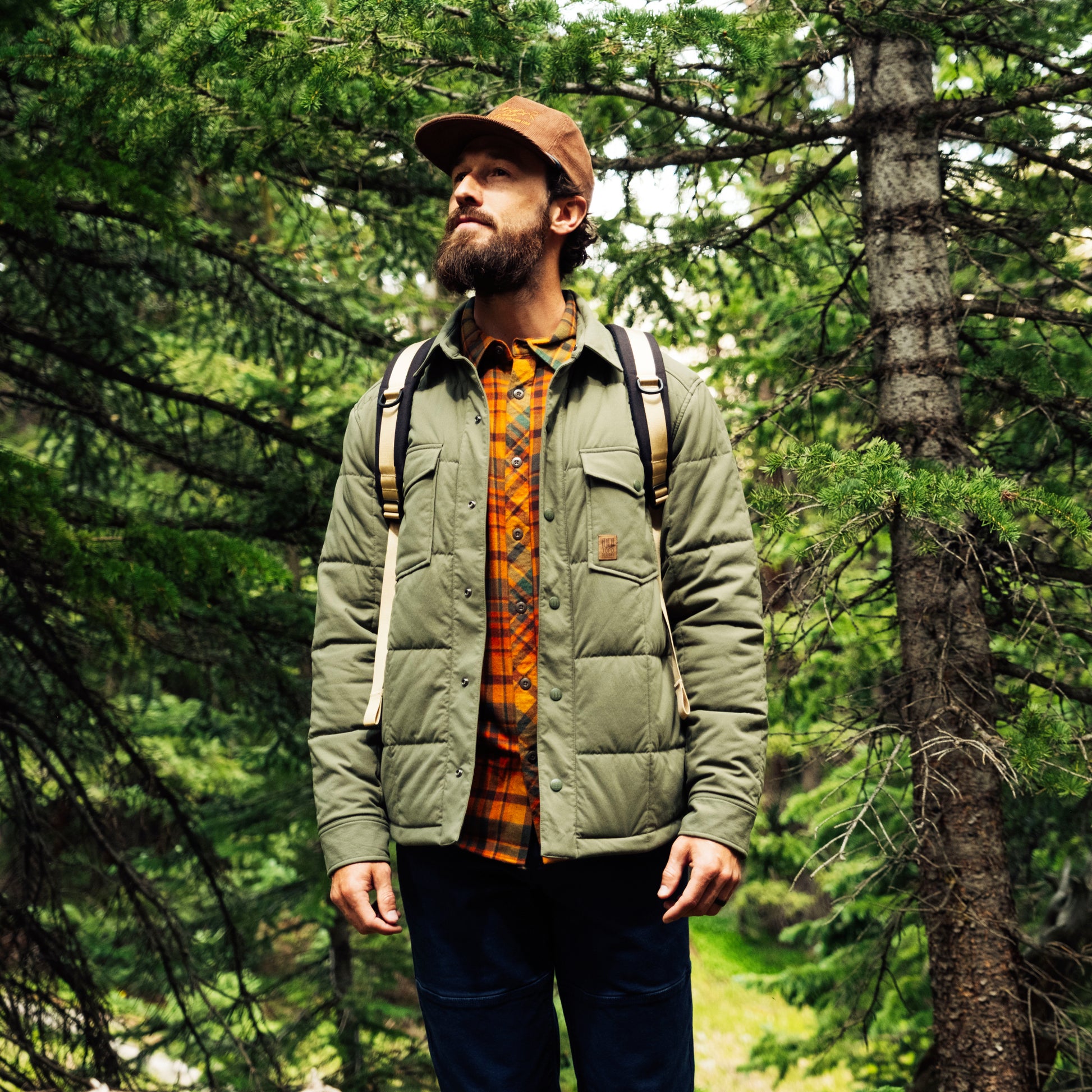 Man in a forest wearing a green jacket and brown cap with a backpack.