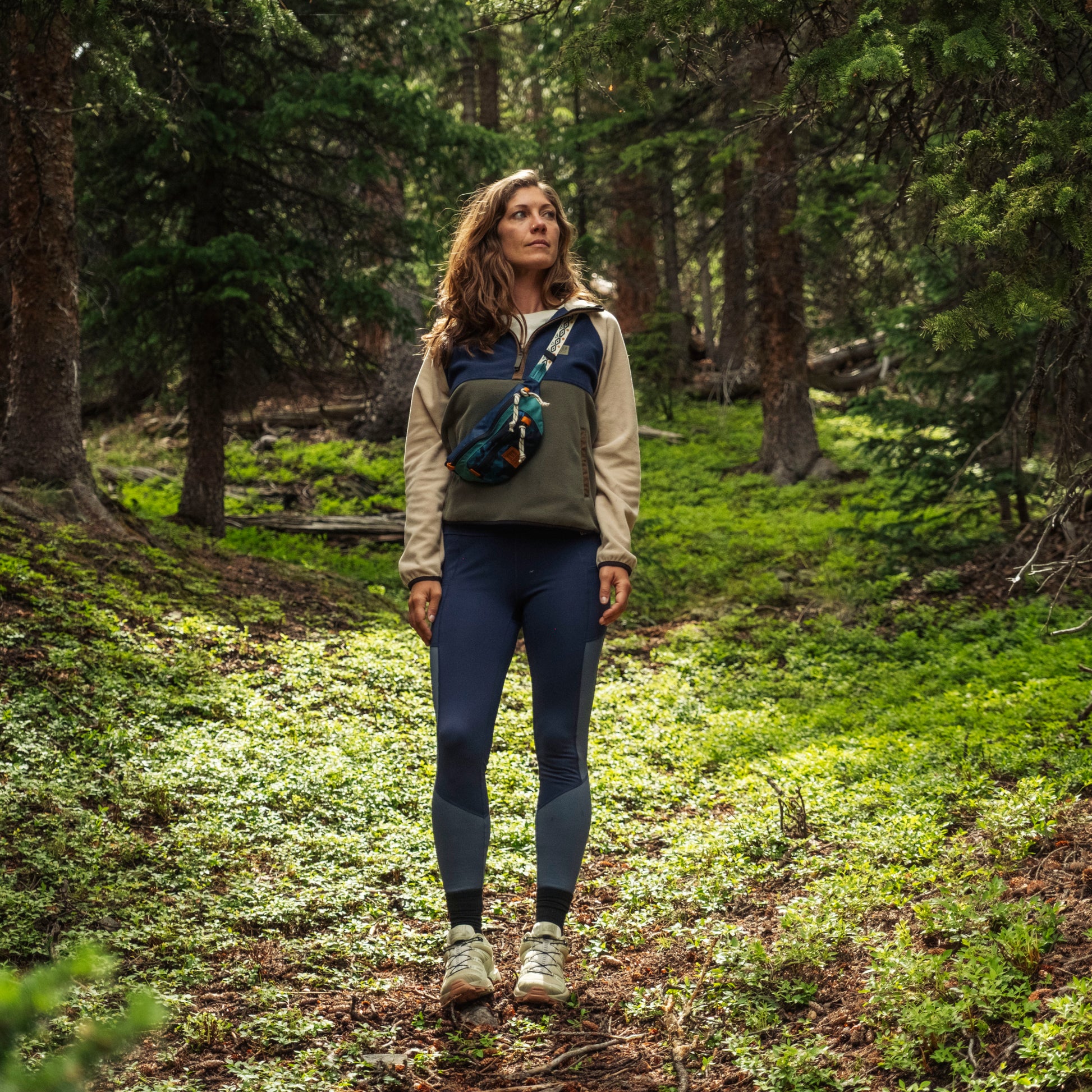 Woman hiking through a forest wearing a green jacket and blue leggings.