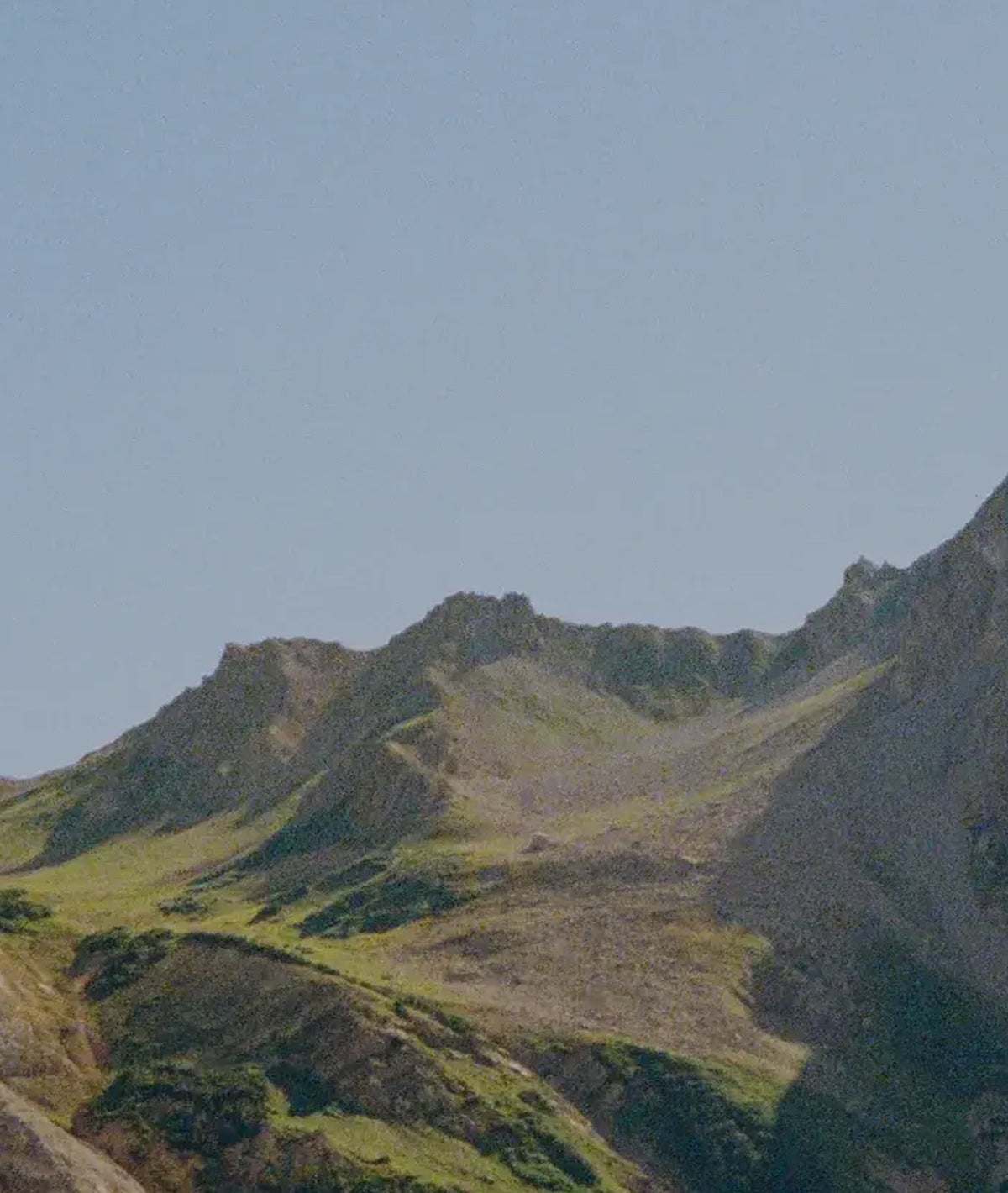A scenic photograph of a rugged mountain ridge with grassy green slopes and rocky gray peaks under a pale blue sky.
