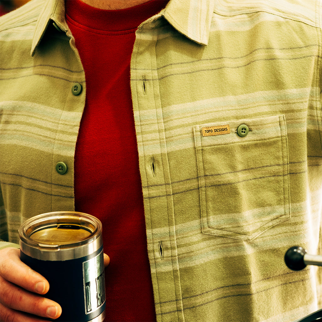 Person wearing a "Oil Green Horizontal Stripe" Mountain Shirt Lightweight with a red sweater underneath, holding a black insulated mug.