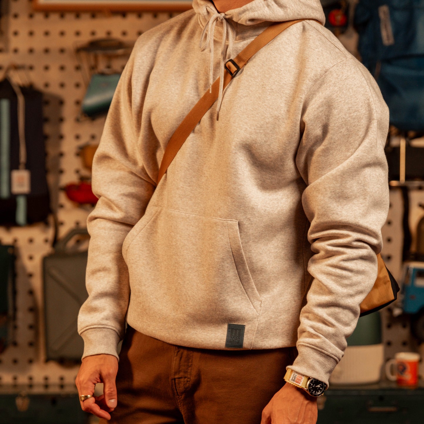 Person wearing a "Oatmeal Heather" hoodie with a brown strap over a pegboard wall.