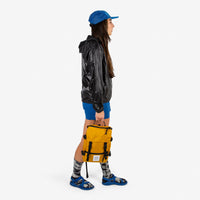 Topo Designs Rover Pack Mini backpack in "Mustard" yellow held by model with top carry handle.